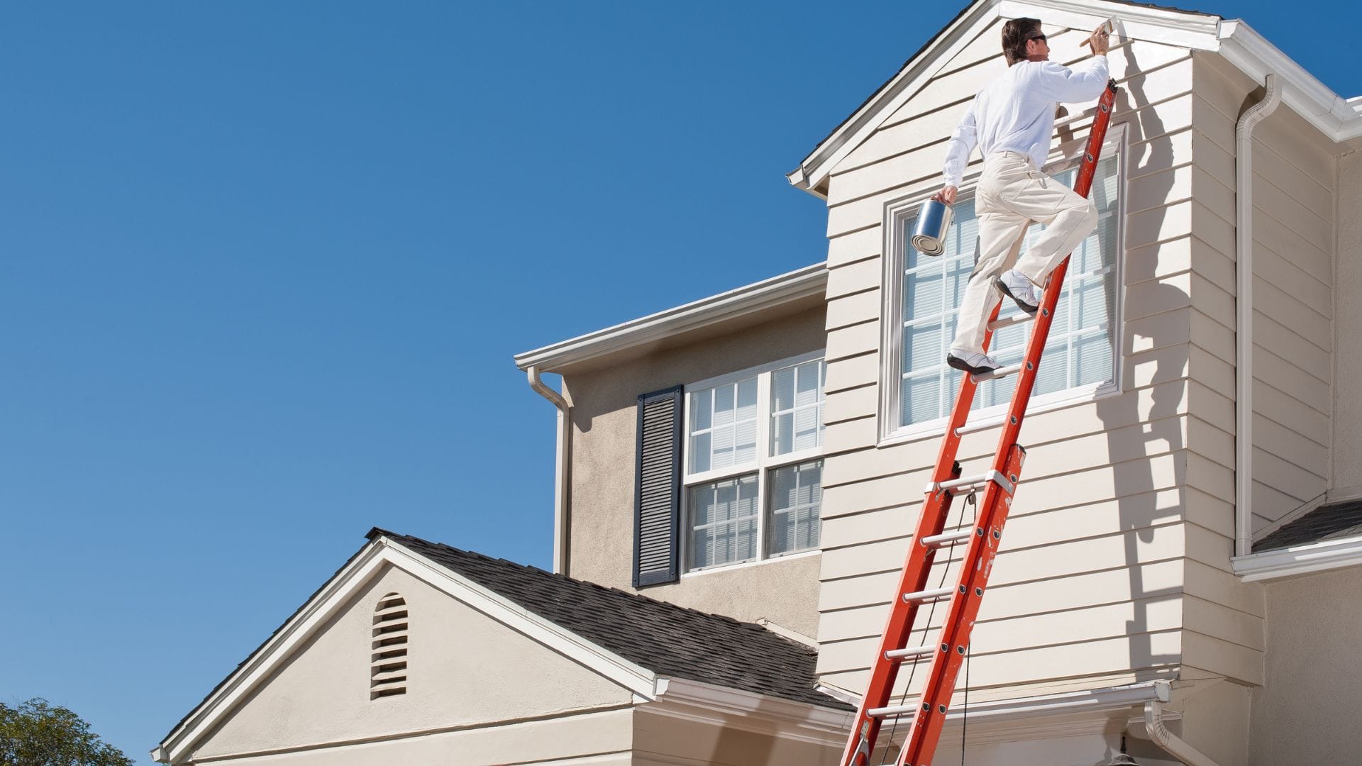 Painter on a ladder brushing the gable trim of a two-story house