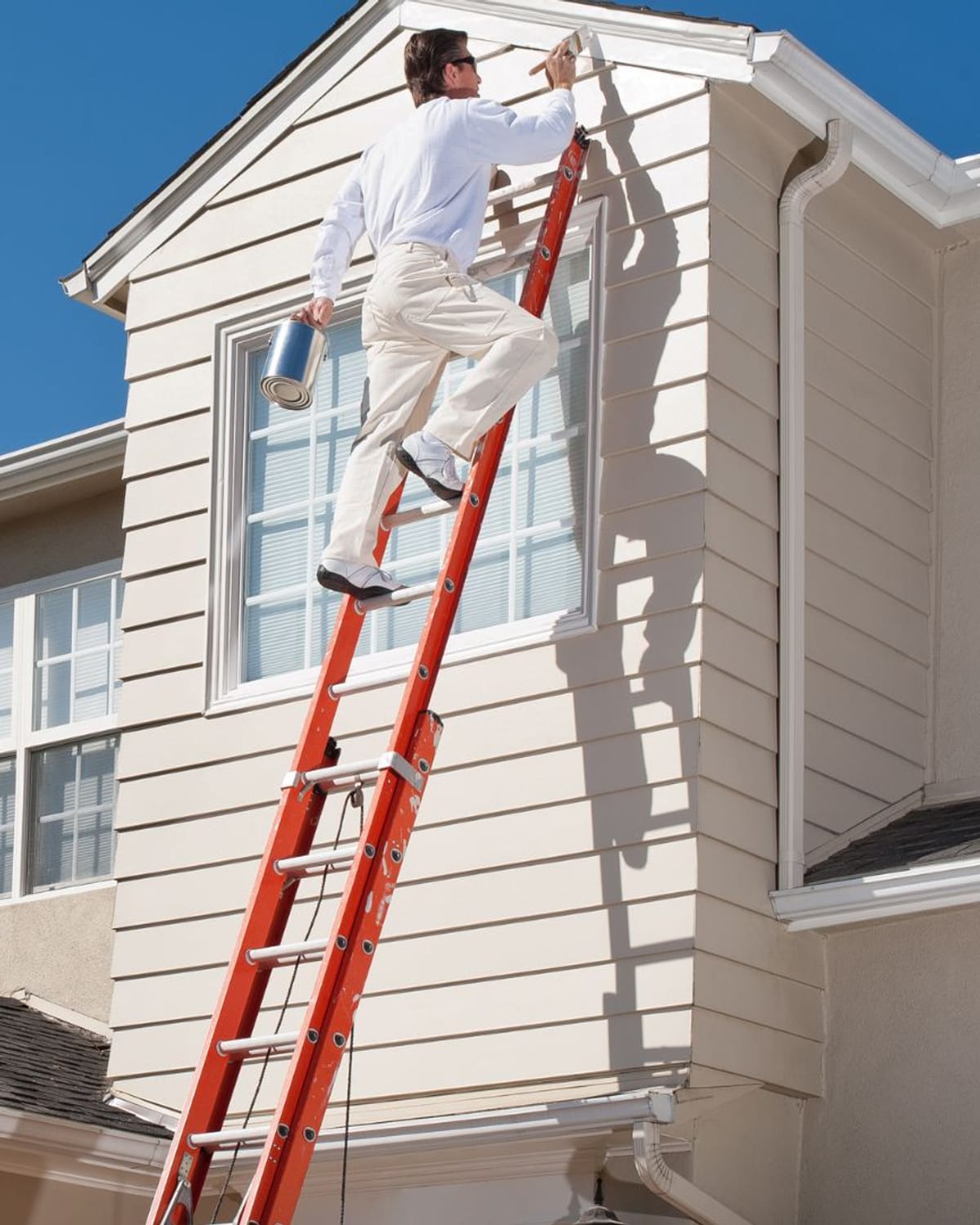 Painter brushing trim from an extension ladder on a two-story home — second-story work adds staging time