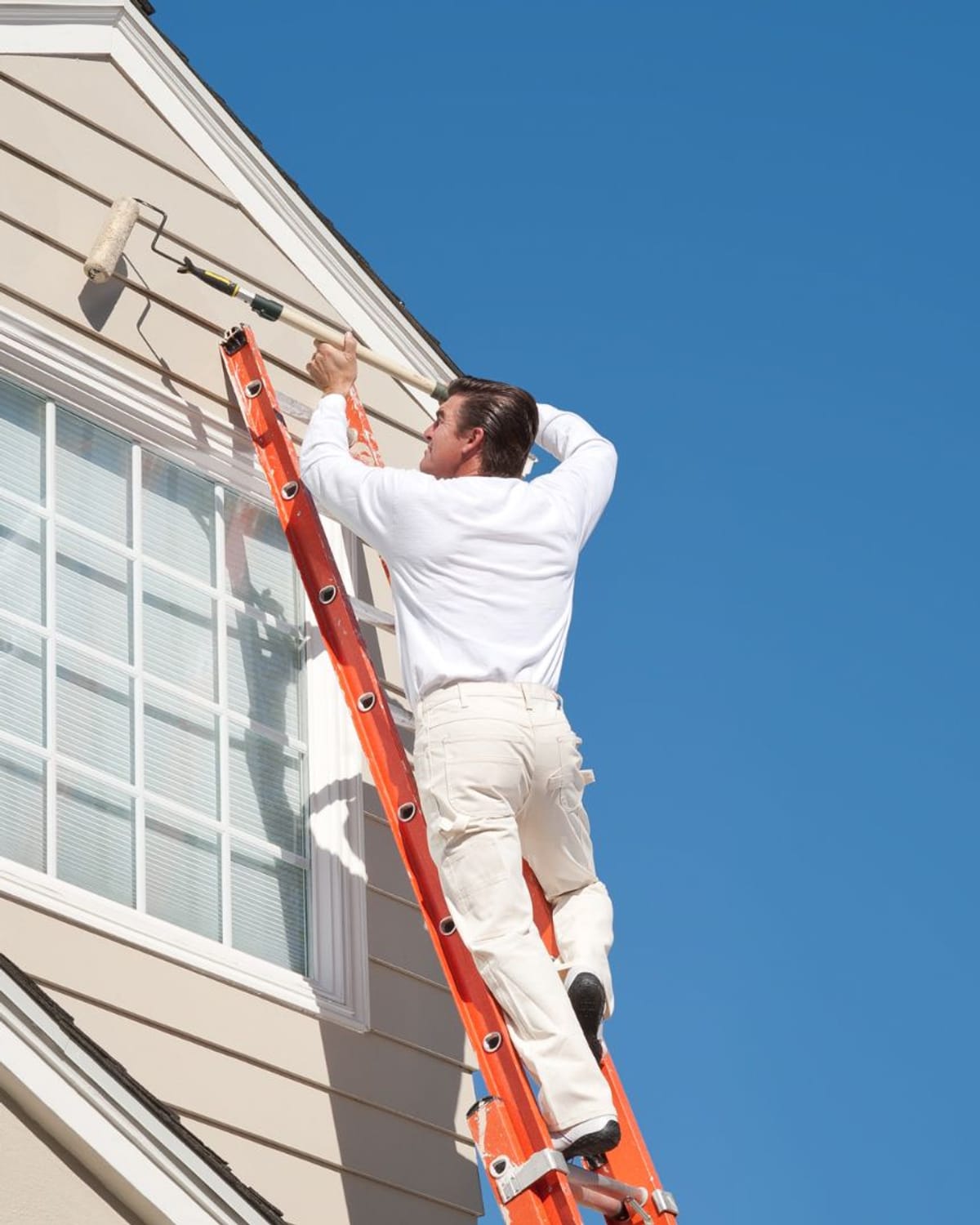 Painter rolling fresh exterior paint onto a gable end from a ladder under a clear Montana sky