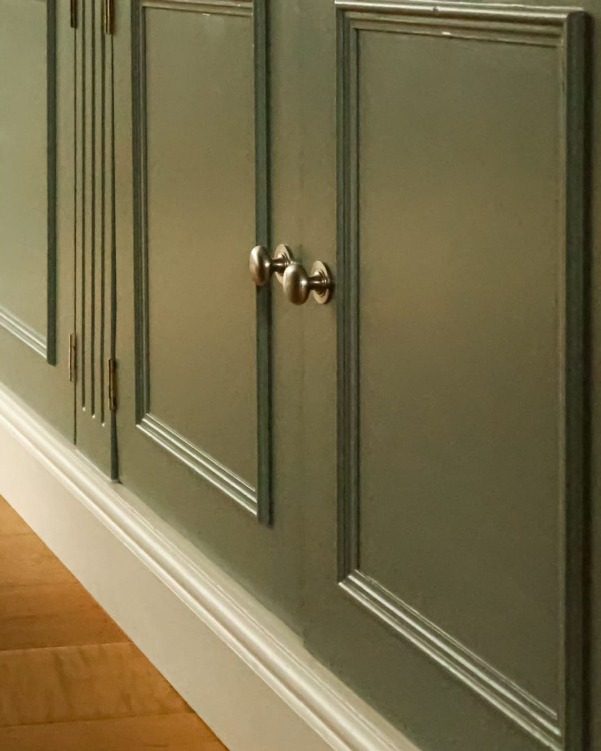 Close-up of sage green painted cabinet doors with brushed nickel knobs and crisp white baseboard